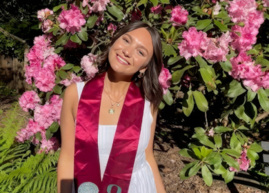 Woman wearing white dress and graduation stole standing in front of flower bush