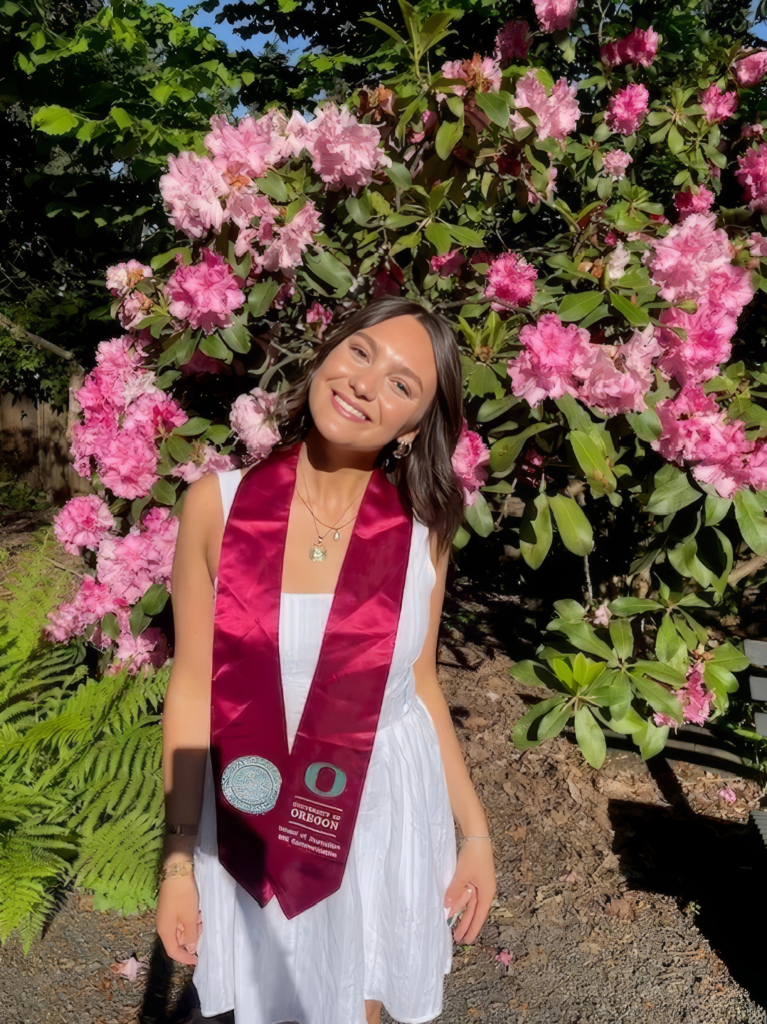 Woman wearing white dress and graduation stole standing in front of flower bush